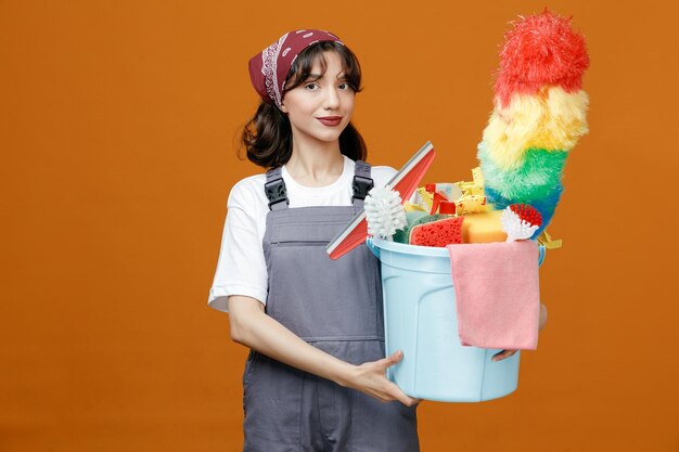 Heureuse jeune femme nettoyante portant l'uniforme et le bandana tenant un seau d'outils de nettoyage avec les deux mains regardant la caméra isolée sur fond orange