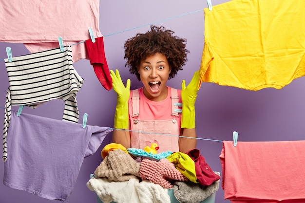 Photo gratuite heureuse jeune femme occupée émotionnelle s'exclame bruyamment, lève les mains dans des gants en caoutchouc, accroche le linge propre sur une corde à linge avec des pinces, a une journée de lavage bien remplie, isolée sur un mur violet. concept d'entretien ménager