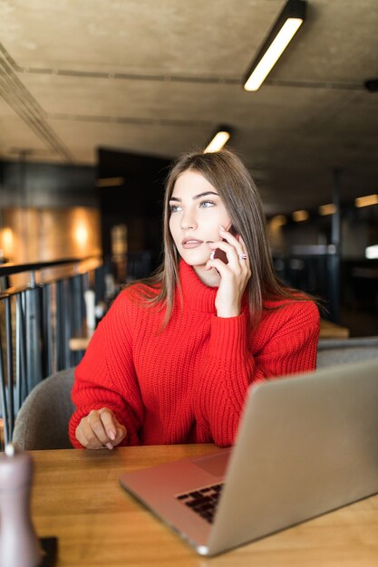 Heureuse jeune femme parlant par téléphone portable et utilisant un ordinateur portable au café