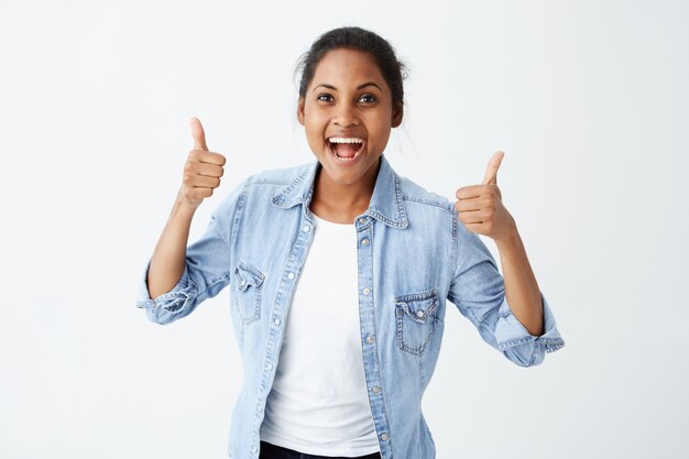 Heureuse jeune femme à la peau foncée portant une chemise à manches longues en jean faisant signe pouce en l'air et souriant joyeusement, montrant son soutien et son respect à quelqu'un. Le langage du corps. Bon travail.