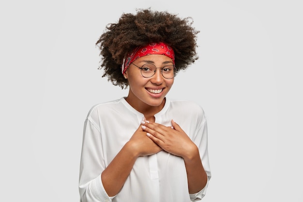 Heureuse jeune femme à la peau sombre et timide avec un large sourire amical garde les mains sur la poitrine, se sent gênée de recevoir le compliment d'un étranger, a les cheveux bouclés et touffus, vêtue d'une chemise blanche à la mode