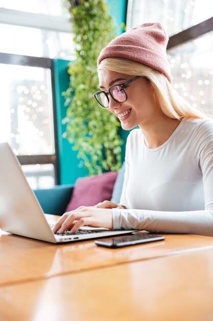 Photo gratuite heureuse jeune femme portant un chapeau et des lunettes à l'aide d'un ordinateur portable