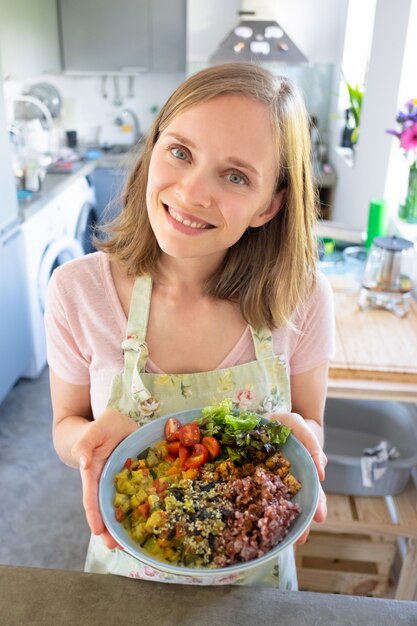 Heureuse jeune femme positive posant avec plat de légumes maison dans sa cuisine, montrant un bol, regardant la caméra et souriant. Tir vertical, grand angle. Concept d'alimentation saine