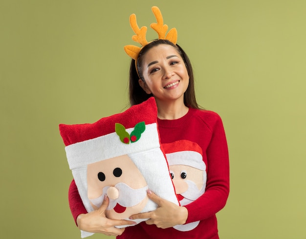 Heureuse jeune femme en pull de Noël rouge portant une jante drôle avec des cornes de cerf tenant un oreiller de Noël à souriant joyeusement