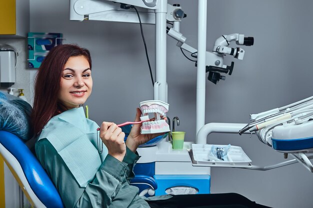 Heureuse jeune femme rousse assise sur une chaise de dentiste et tenant une brosse à dents et un dentier dans un cabinet de dentiste.