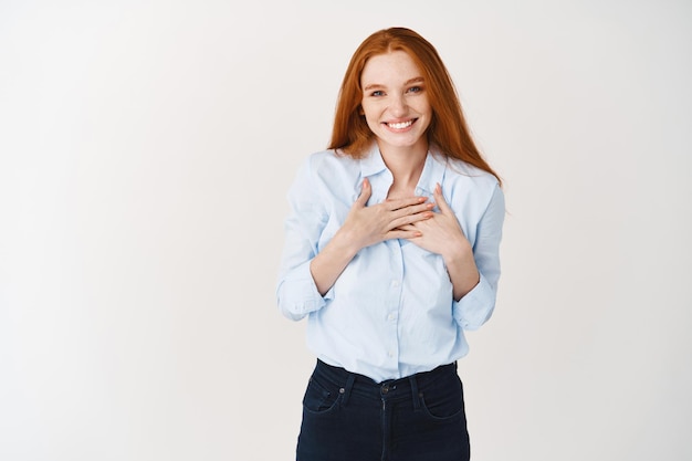 Heureuse jeune femme rousse vous remerciant, tenant les mains sur la poitrine et souriante, l'air reconnaissante devant, mur blanc