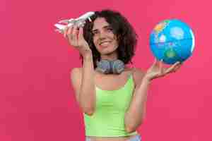 Photo gratuite une heureuse jeune femme séduisante aux cheveux courts en vert crop top dans les écouteurs tenant globe et regardant avion jouet