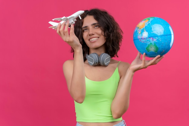 Une heureuse jeune femme séduisante aux cheveux courts en vert crop top dans les écouteurs tenant globe et regardant avion jouet