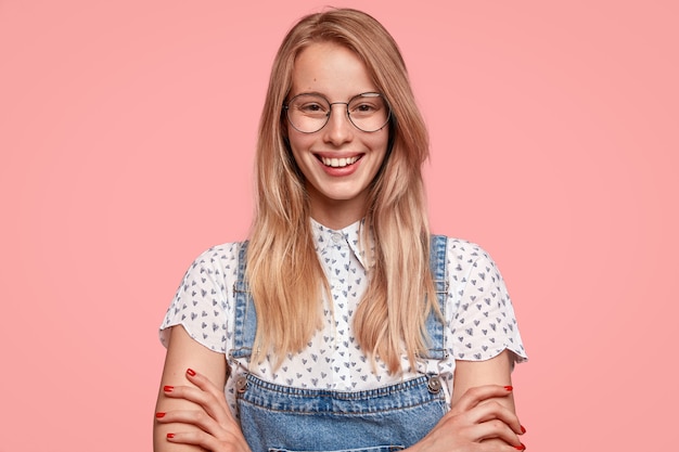 Photo gratuite heureuse jeune femme avec un sourire charmimg, porte une chemise et une salopette élégantes, garde les mains croisées, se réjouit des moments positifs de la vie, heureuse de recevoir un bonus du patron pour un travail diligent