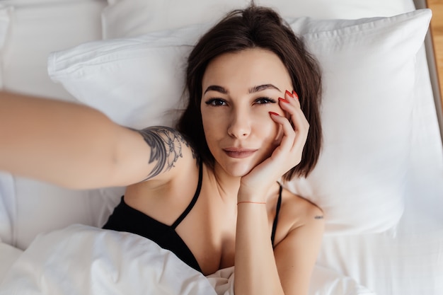 Heureuse jeune femme tenir un téléphone intelligent couché éveillé dans son lit le matin faisant vue de dessus selfie