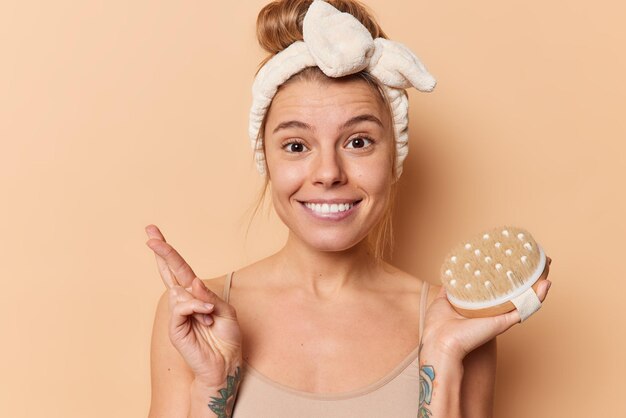 Heureuse jeune femme tient une brosse pour le corps garde les doigts croisés espère bonne chance subit des procédures de beauté et de traitement de la peau prend soin d'elle-même porte un bandeau t-shirt décontracté pose à l'intérieur