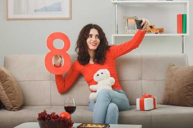 Heureuse jeune femme en vêtements décontractés assis sur un canapé avec présent tenant le numéro huit et ours en peluche faisant selfie à l'aide de smartphone souriant célébrant la journée internationale de la femme le 8 mars