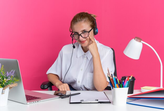 Heureuse jeune fille blonde du centre d'appels portant un casque et des lunettes assise au bureau avec des outils de travail pointant le doigt sur la joue en regardant la caméra isolée sur le mur rose