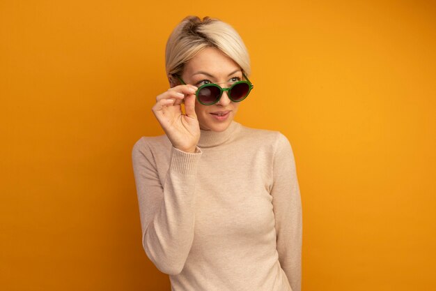 Heureuse jeune fille blonde portant et saisissant des lunettes de soleil regardant le côté isolé sur un mur orange avec espace de copie