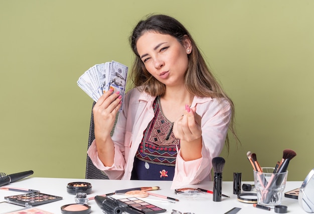 Heureuse jeune fille brune assise à table avec des outils de maquillage tenant de l'argent et gesticulant un signe d'argent