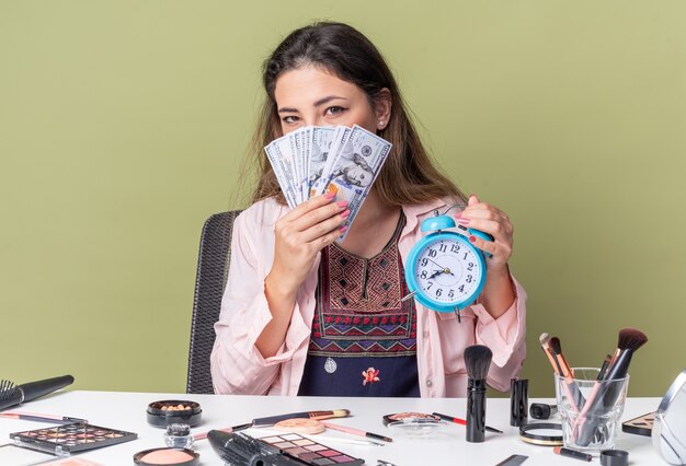 Heureuse jeune fille brune assise à table avec des outils de maquillage tenant de l'argent et un réveil