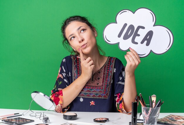 Heureuse jeune fille brune assise à table avec des outils de maquillage tenant une bulle d'idée et un pinceau de maquillage en levant