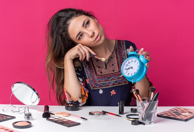 Heureuse jeune fille brune assise à table avec des outils de maquillage tenant un réveil et un pinceau de maquillage