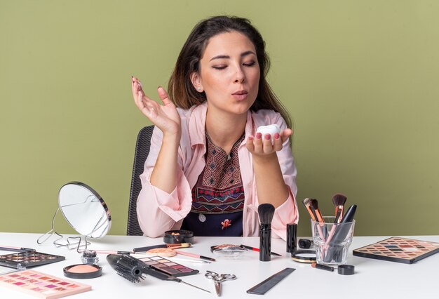 Heureuse jeune fille brune assise à table avec des outils de maquillage tenant et soufflant sur une mousse pour cheveux isolée sur un mur vert olive avec espace de copie