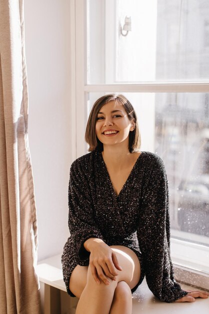 Heureuse jeune fille brune caucasienne en robe noire à la mode souriant à la caméra tout en étant assis sur le rebord de la fenêtre Concept de bonne humeur