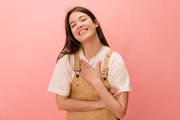 Heureuse jeune fille brune caucasienne tenant la main sur la poitrine souriant à la caméra sur fond rose Concept d'émotions et d'états d'esprit