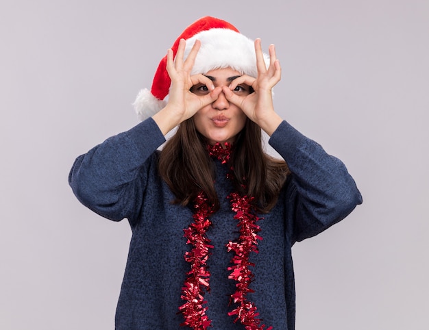 Heureuse jeune fille caucasienne avec bonnet de noel et guirlande autour du cou à travers les doigts isolés sur un mur blanc avec espace de copie