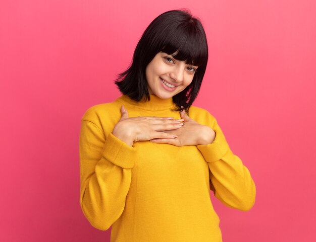 Heureuse jeune fille caucasienne brune met les mains sur la poitrine et regarde la caméra