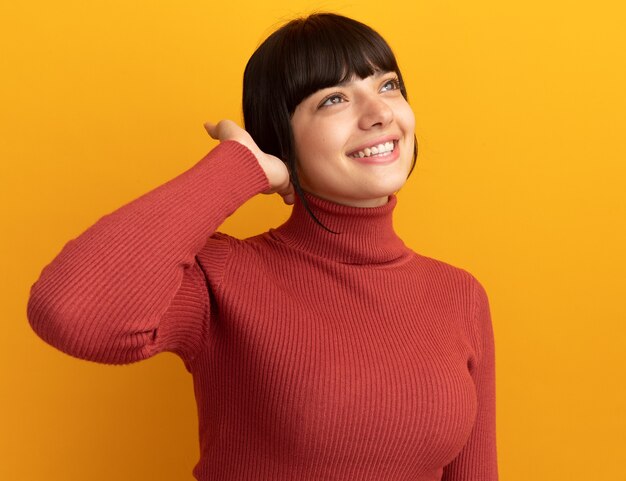 Heureuse jeune fille caucasienne brune tient la main près de la tête et lève les yeux