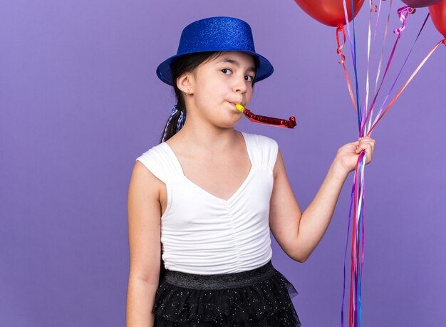 heureuse jeune fille caucasienne avec un chapeau de fête bleu tenant des ballons à l'hélium et soufflant un sifflet de fête isolé sur un mur violet avec espace pour copie