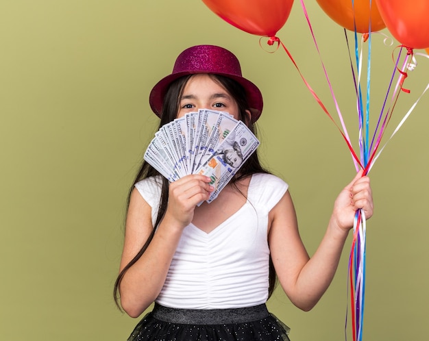 heureuse jeune fille caucasienne avec un chapeau de fête violet tenant des ballons à l'hélium et de l'argent devant son visage isolé sur un mur vert olive avec espace pour copie