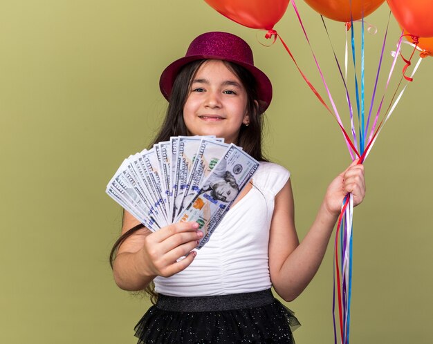 heureuse jeune fille caucasienne avec chapeau de fête violet tenant des ballons à l'hélium et de l'argent isolé sur un mur vert olive avec espace de copie