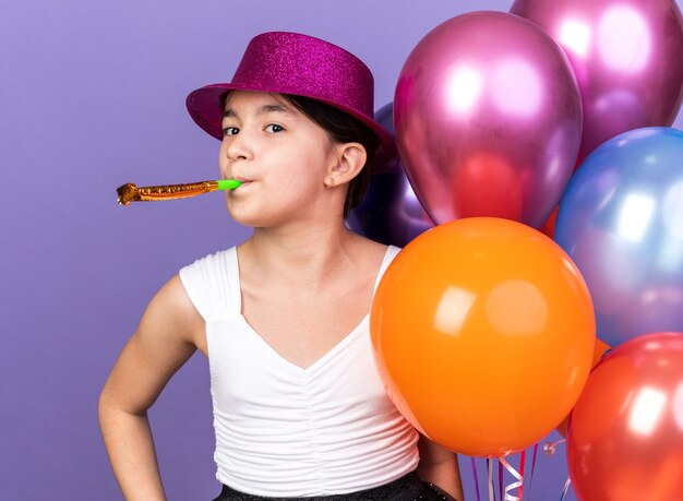 heureuse jeune fille caucasienne avec un chapeau de fête violet tenant des ballons à l'hélium et soufflant un sifflet de fête isolé sur un mur violet avec espace de copie