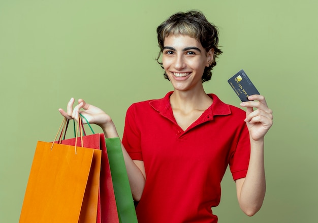 Heureuse jeune fille caucasienne avec coupe de cheveux de lutin tenant des sacs à provisions et une carte de crédit sur un espace vert olive