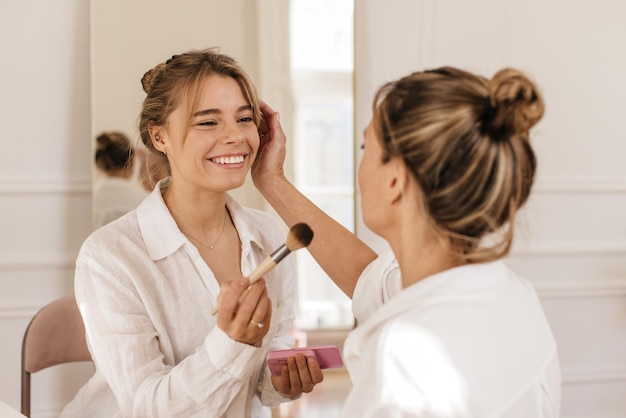 Heureuse jeune fille caucasienne avec son amie, apprenez à vous maquiller, commencez par appliquer de la poudre. Concept de procédure de beauté