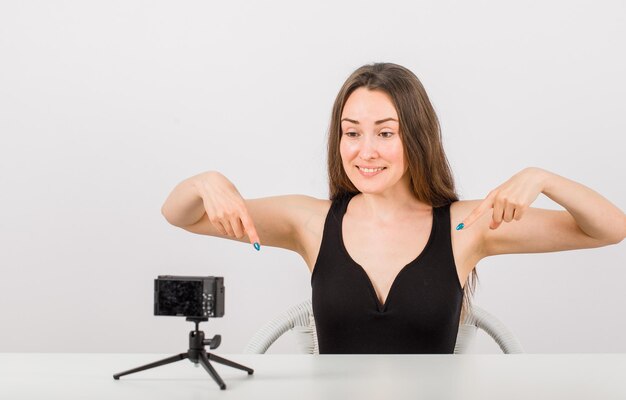 Heureuse jeune fille pose à la petite caméra en pointant vers le bas avec les index sur fond blanc