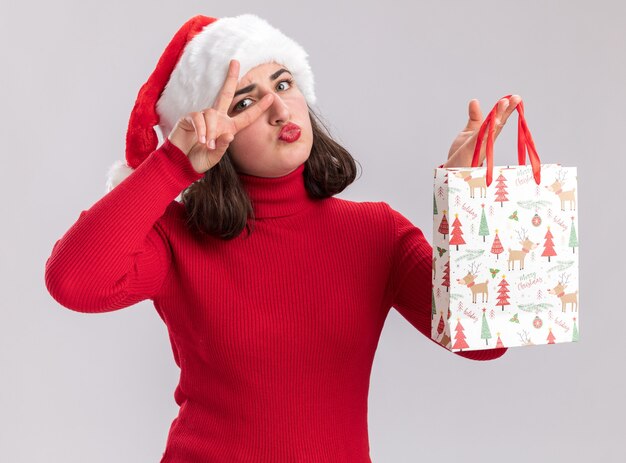 Heureuse jeune fille en pull rouge et bonnet de Noel tenant un sac en papier coloré avec des cadeaux de Noël montrant v-sign près des yeux debout sur un mur blanc