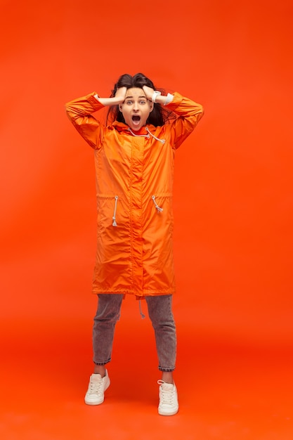 L'heureuse Jeune Fille Souriante Posant Au Studio En Veste Orange Automne Isolé Sur Rouge. émotions Positives Humaines. Concept Du Temps Froid. Concepts De Mode Féminine