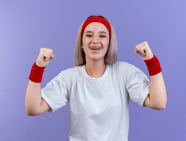 Heureuse jeune fille sportive caucasienne avec des bretelles portant un bandeau et des bracelets garde les poings