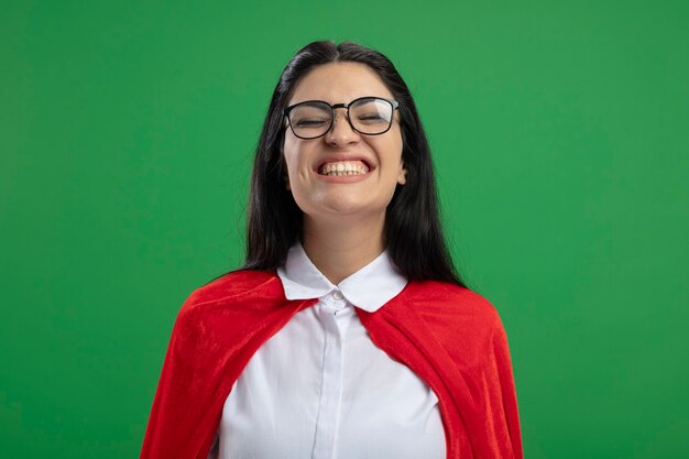 Heureuse jeune fille de super-héros caucasien portant des lunettes souriant sans aucun mouvement avec les yeux fermés isolé sur un mur vert avec espace copie