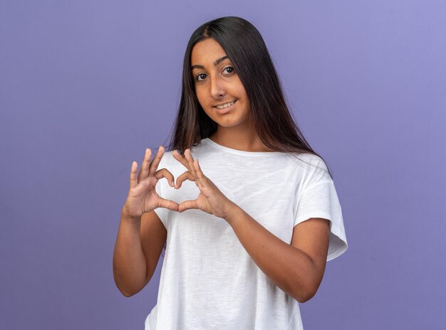 Heureuse jeune fille en t-shirt blanc regardant la caméra en souriant faisant un geste du cœur avec les doigts debout sur fond bleu