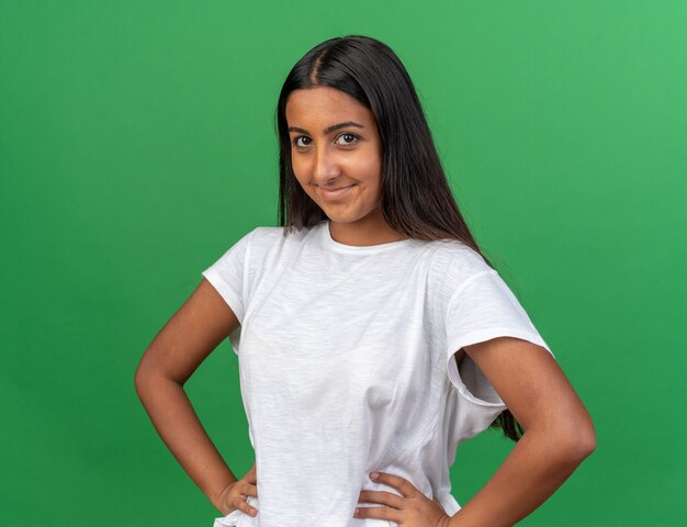 Heureuse jeune fille en t-shirt blanc regardant la caméra souriante confiante avec les bras sur la hanche debout sur fond vert