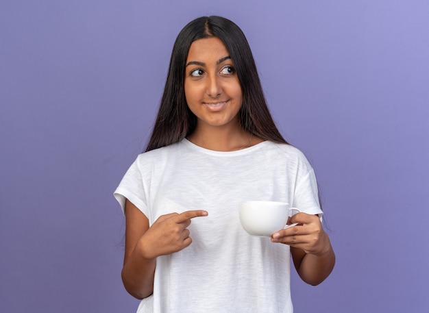 Heureuse jeune fille en t-shirt blanc tenant une tasse blanche pointant avec l'index en regardant de côté avec le sourire sur le visage debout sur fond bleu