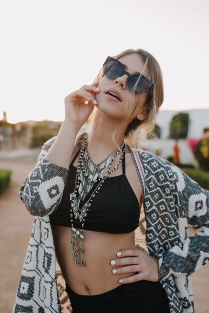 Heureuse jeune fille en vacances, tirez sur la plage. Porter un maillot de bain noir, des lunettes de soleil, un collier élégant, un cardigan. Profiter du coucher du soleil, des vacances, de l'été