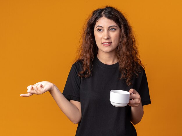Heureuse jeune jolie femme tenant une tasse de thé regardant et pointant sur le côté isolé sur un mur orange