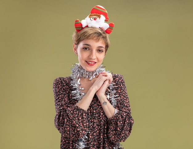 Photo gratuite heureuse jeune jolie fille portant un bandeau de père noël et une guirlande de guirlandes autour du cou en gardant les mains ensemble isolées sur un mur vert olive avec espace de copie