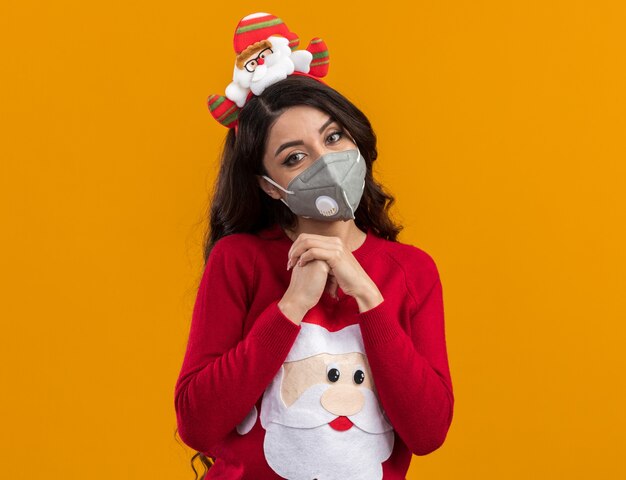 Heureuse jeune jolie fille portant un bandeau de père noël et un pull avec un masque de protection gardant les mains ensemble isolées sur un mur orange avec espace de copie