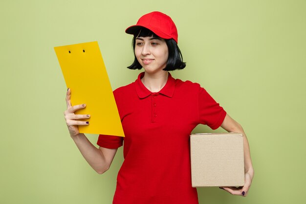 Heureuse jeune livreuse caucasienne tenant une boîte en carton et regardant le presse-papiers