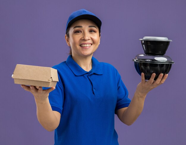 Heureuse jeune livreuse en uniforme bleu et casquette tenant des colis alimentaires souriant joyeusement