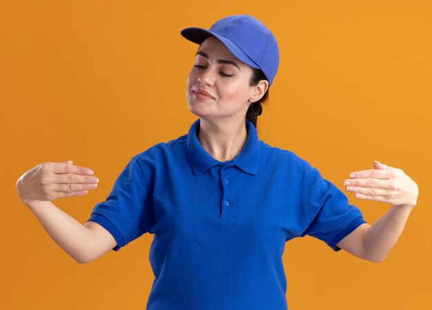 Heureuse jeune livreuse en uniforme et casquette faisant semblant de tenir quelque chose devant elle en le regardant isolé sur un mur orange