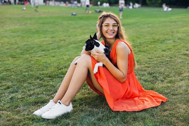 Heureuse jolie femme assise sur l'herbe dans le parc d'été, tenant un chien Boston Terrier, souriant humeur positive, vêtue d'une robe orange, style branché, jambes minces, baskets, jouant avec un animal de compagnie